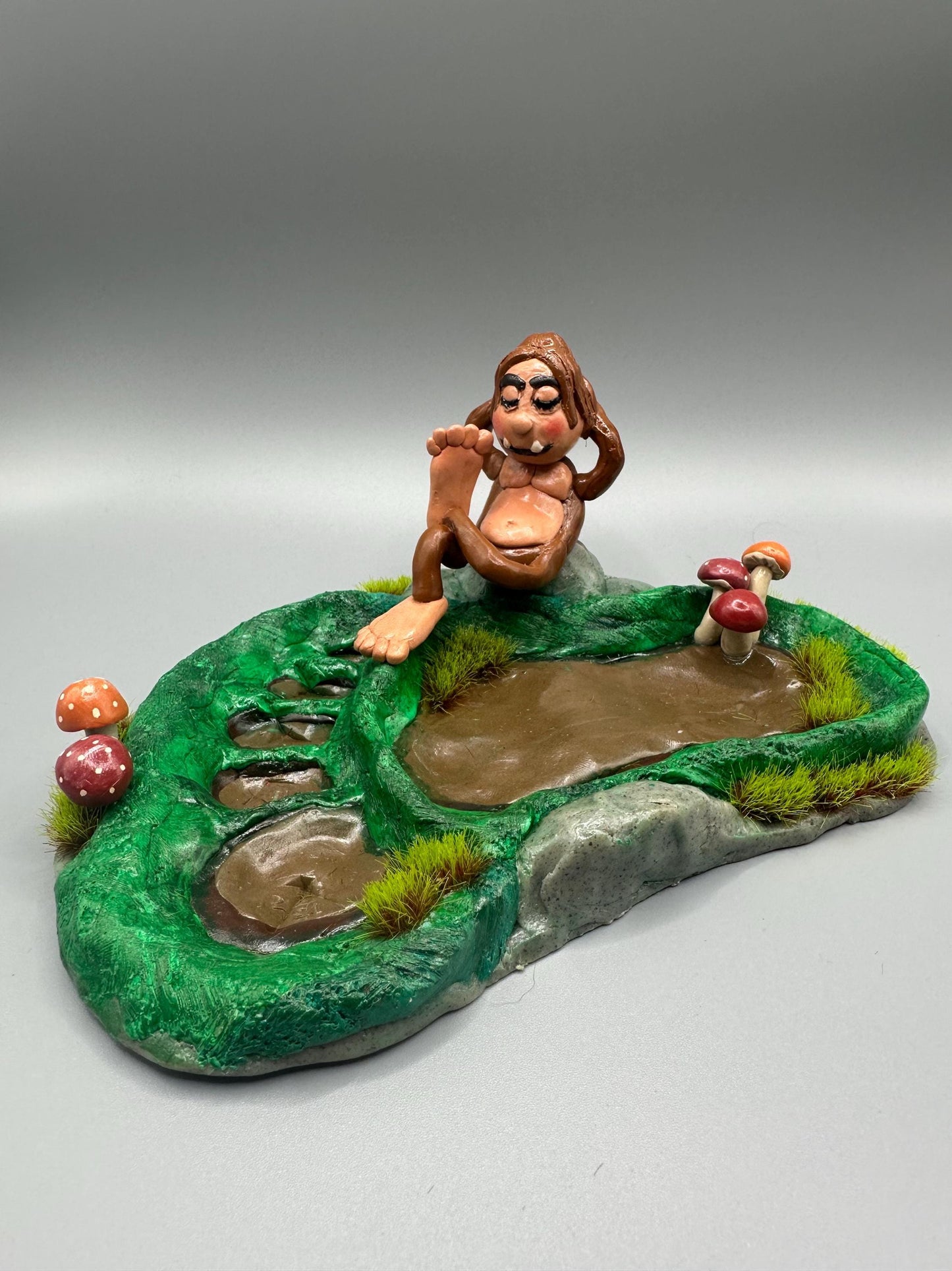 Cute handmade polymer clay dice tray and holder for a cryptid lover with a baby Bigfoot theme. Perfect gift for RPG, D&D, cryptid lovers and fantasy lovers. Baby Bigfoot sitting at the edge of a full grown Bigfoot foot print in the woods surrounded by fake grass and clay mushrooms.