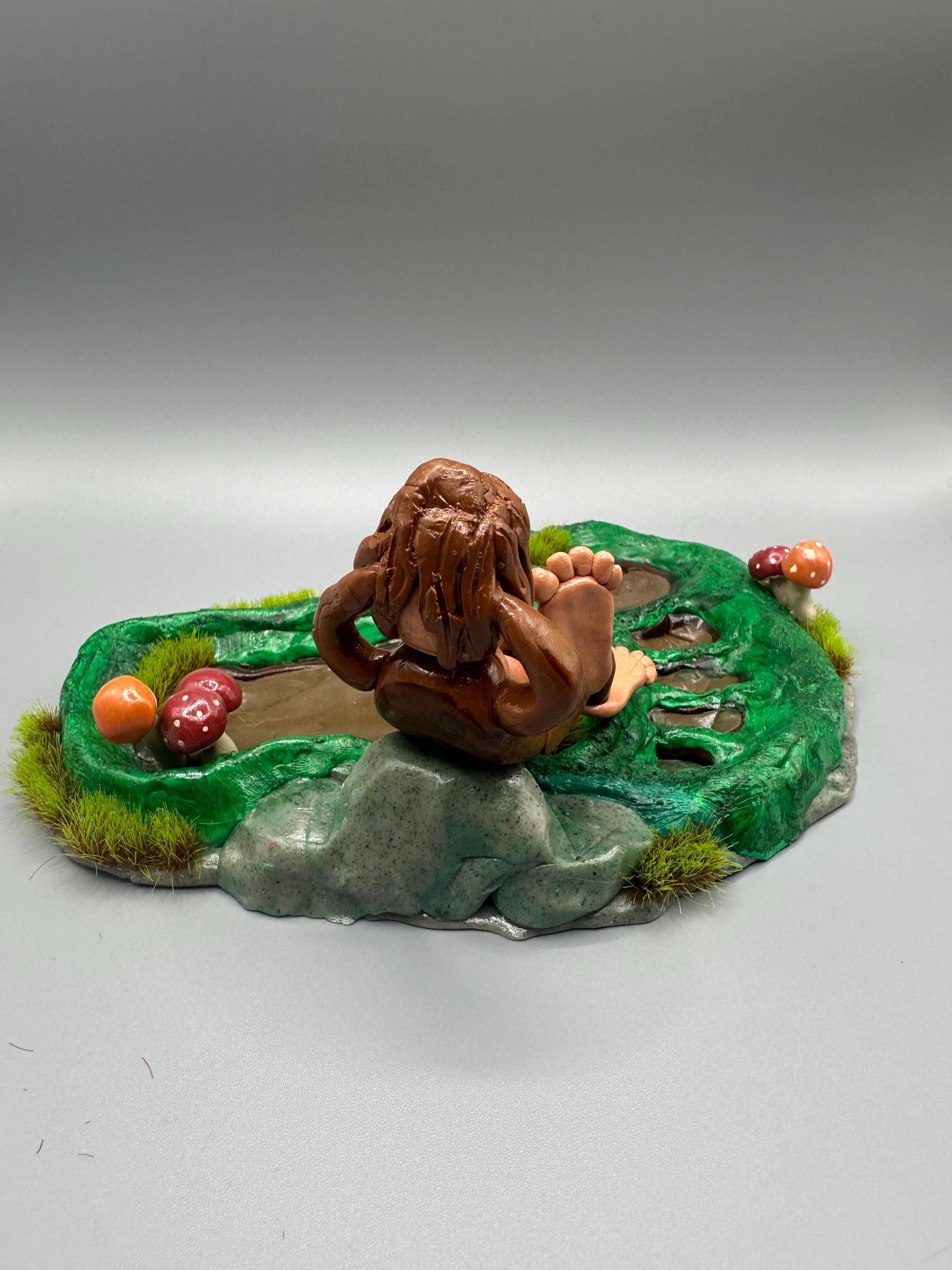Cute handmade polymer clay dice tray and holder for a cryptid lover with a baby Bigfoot theme. Perfect gift for RPG, D&D, cryptid lovers and fantasy lovers. Baby Bigfoot sitting at the edge of a full grown Bigfoot foot print in the woods surrounded by fake grass and clay mushrooms.
