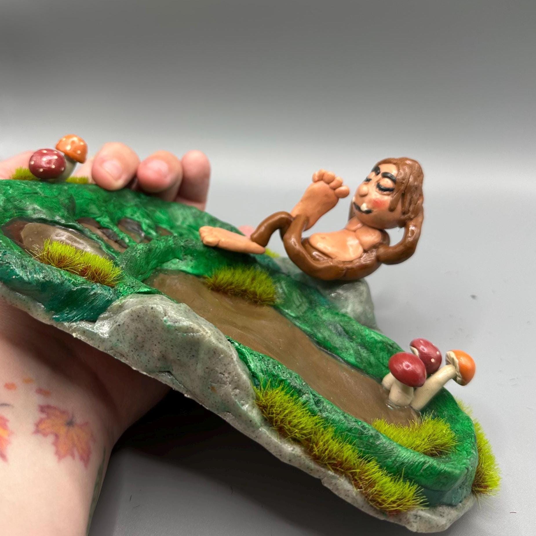 Cute handmade polymer clay dice tray and holder for a cryptid lover with a baby Bigfoot theme. Perfect gift for RPG, D&D, cryptid lovers and fantasy lovers. Baby Bigfoot sitting at the edge of a full grown Bigfoot foot print in the woods surrounded by fake grass and clay mushrooms.