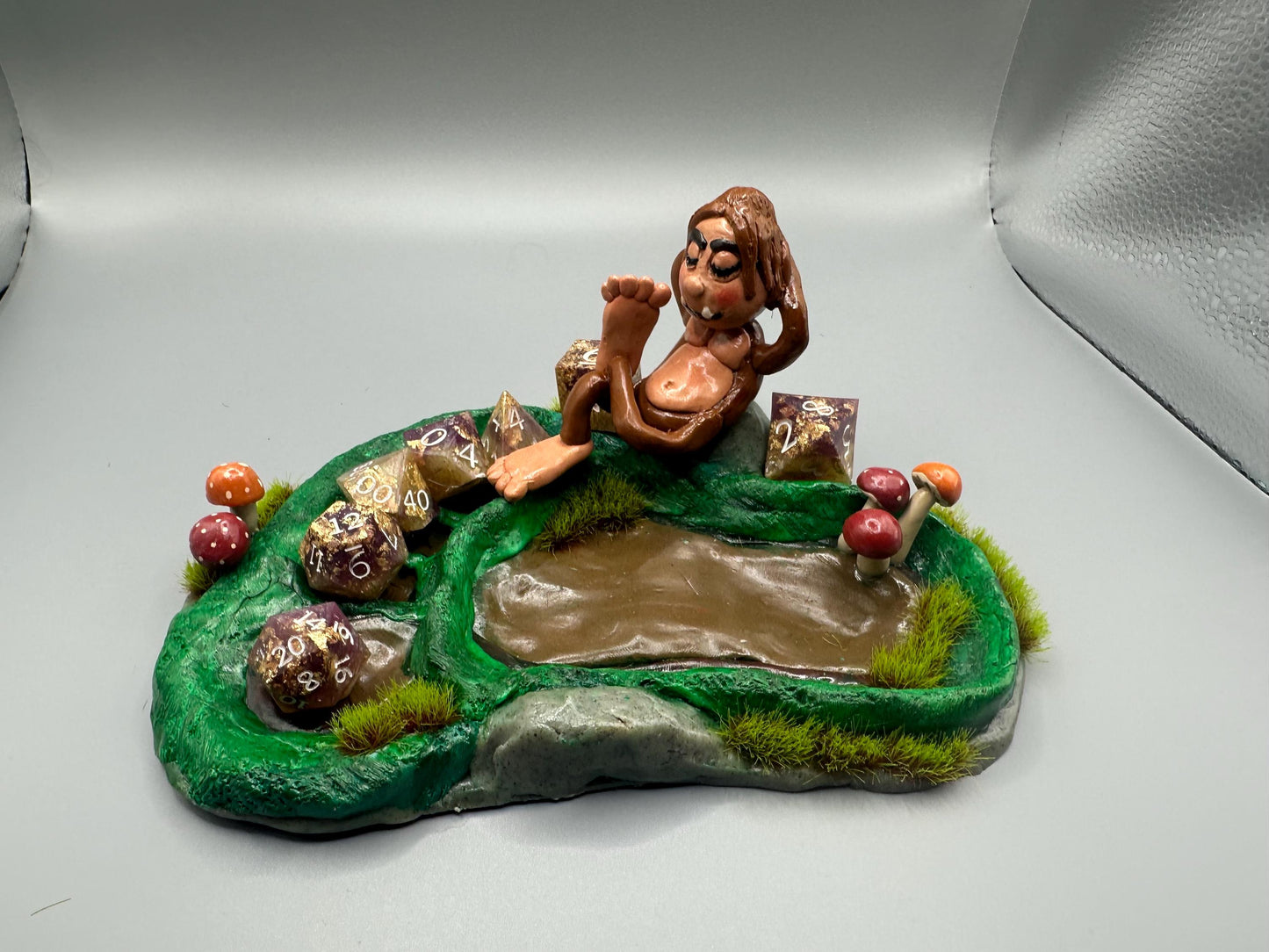 Cute handmade polymer clay dice tray and holder for a cryptid lover with a baby Bigfoot theme. Perfect gift for RPG, D&D, cryptid lovers and fantasy lovers. Baby Bigfoot sitting at the edge of a full grown Bigfoot foot print in the woods surrounded by fake grass and clay mushrooms. No AI used in the making of this dice tray!