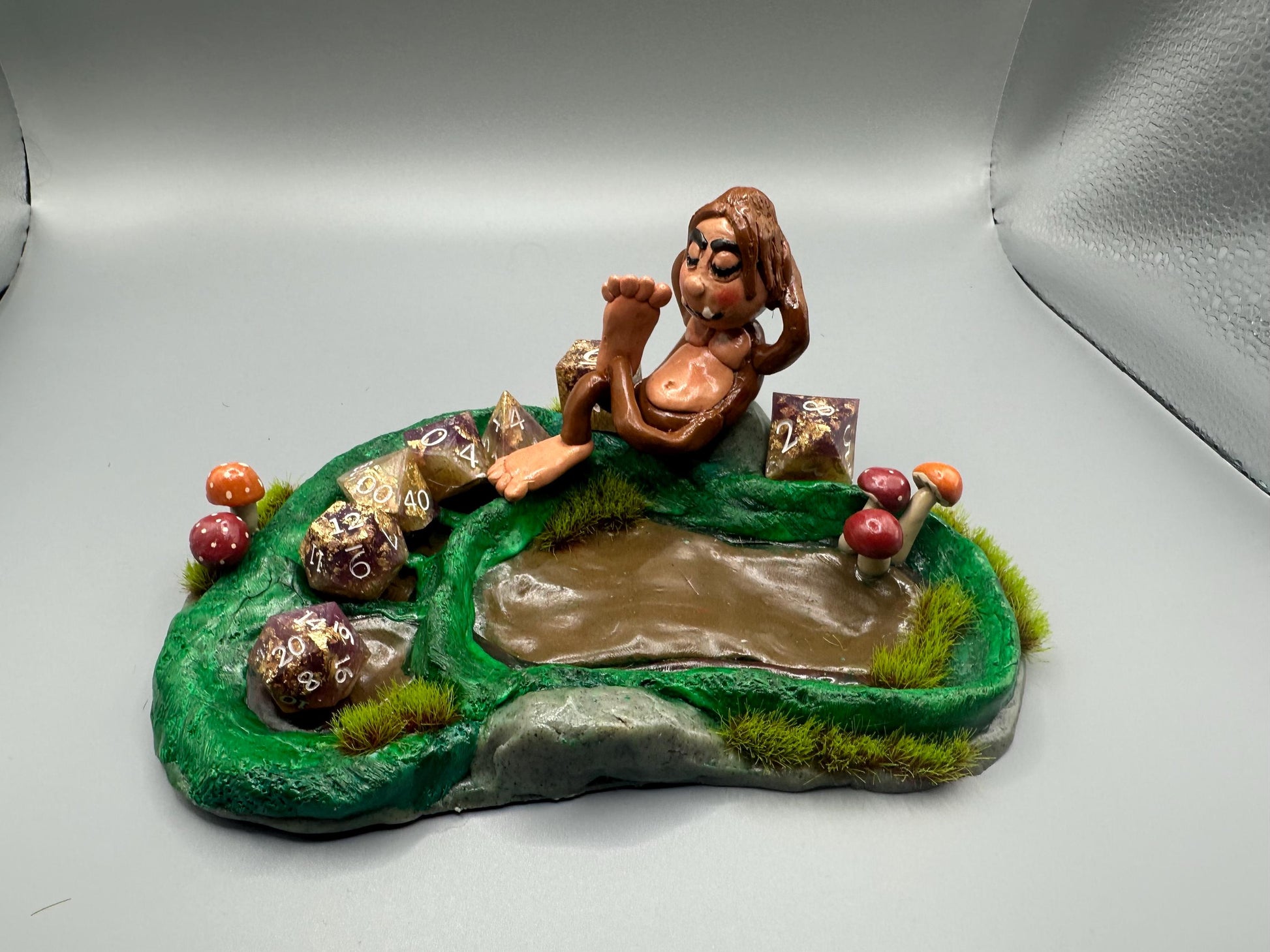 Cute handmade polymer clay dice tray and holder for a cryptid lover with a baby Bigfoot theme. Perfect gift for RPG, D&D, cryptid lovers and fantasy lovers. Baby Bigfoot sitting at the edge of a full grown Bigfoot foot print in the woods surrounded by fake grass and clay mushrooms. No AI used in the making of this dice tray!