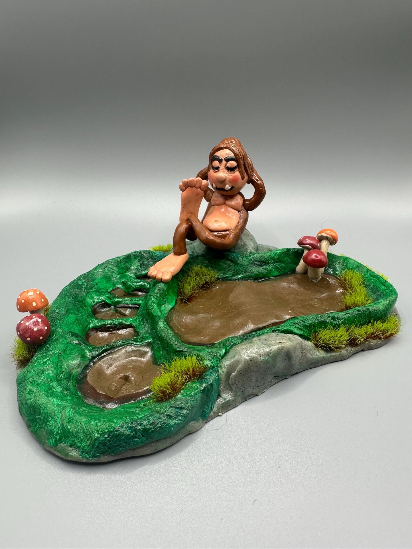 Cute handmade polymer clay dice tray and holder for a cryptid lover with a baby Bigfoot theme. Perfect gift for RPG, D&D, cryptid lovers and fantasy lovers. Baby Bigfoot sitting at the edge of a full grown Bigfoot foot print in the woods surrounded by fake grass and clay mushrooms. No AI used in the making of this dice tray!
