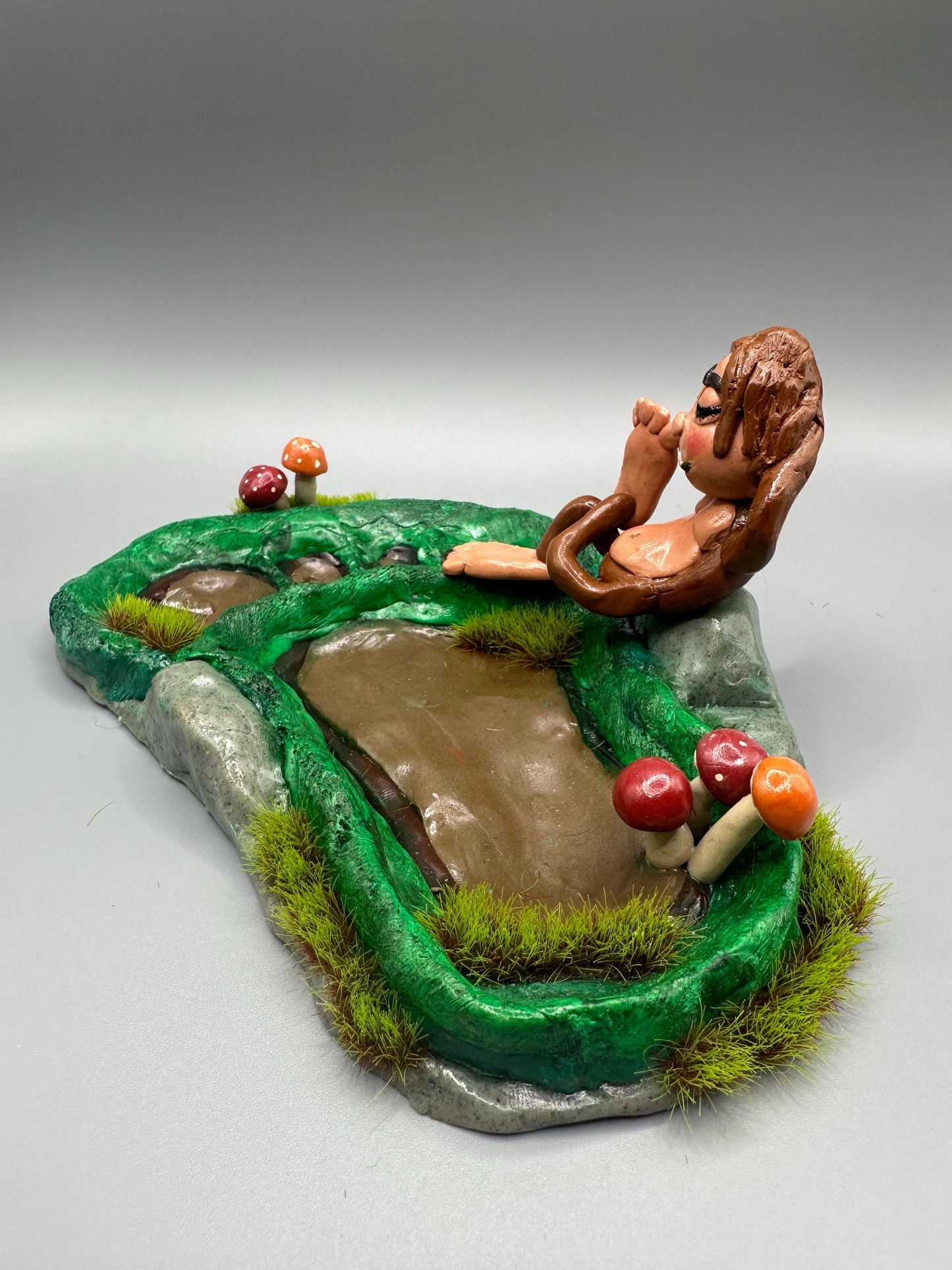 Cute handmade polymer clay dice tray and holder for a cryptid lover with a baby Bigfoot theme. Perfect gift for RPG, D&D, cryptid lovers and fantasy lovers. Baby Bigfoot sitting at the edge of a full grown Bigfoot foot print in the woods surrounded by fake grass and clay mushrooms. No AI used in the making of this dice tray!