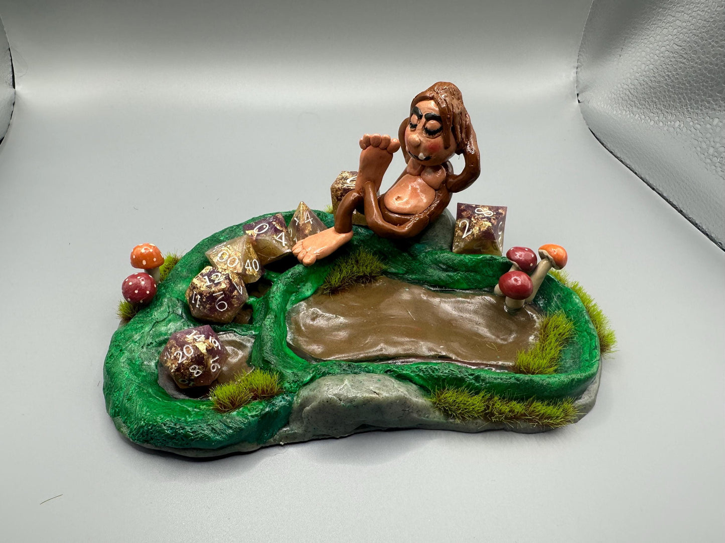 Cute handmade polymer clay dice tray and holder for a cryptid lover with a baby Bigfoot theme. Perfect gift for RPG, D&D, cryptid lovers and fantasy lovers. Baby Bigfoot sitting at the edge of a full grown Bigfoot foot print in the woods surrounded by fake grass and clay mushrooms. No AI used in the making of this dice tray!