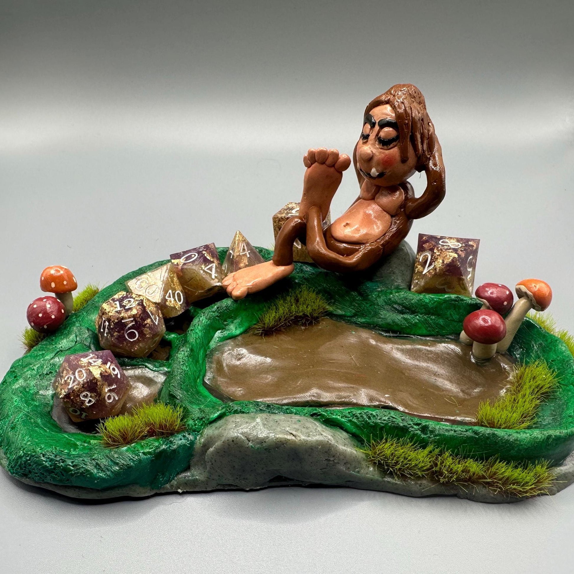 Cute handmade polymer clay dice tray and holder for a cryptid lover with a baby Bigfoot theme. Perfect gift for RPG, D&D, cryptid lovers and fantasy lovers. Baby Bigfoot sitting at the edge of a full grown Bigfoot foot print in the woods surrounded by fake grass and clay mushrooms.