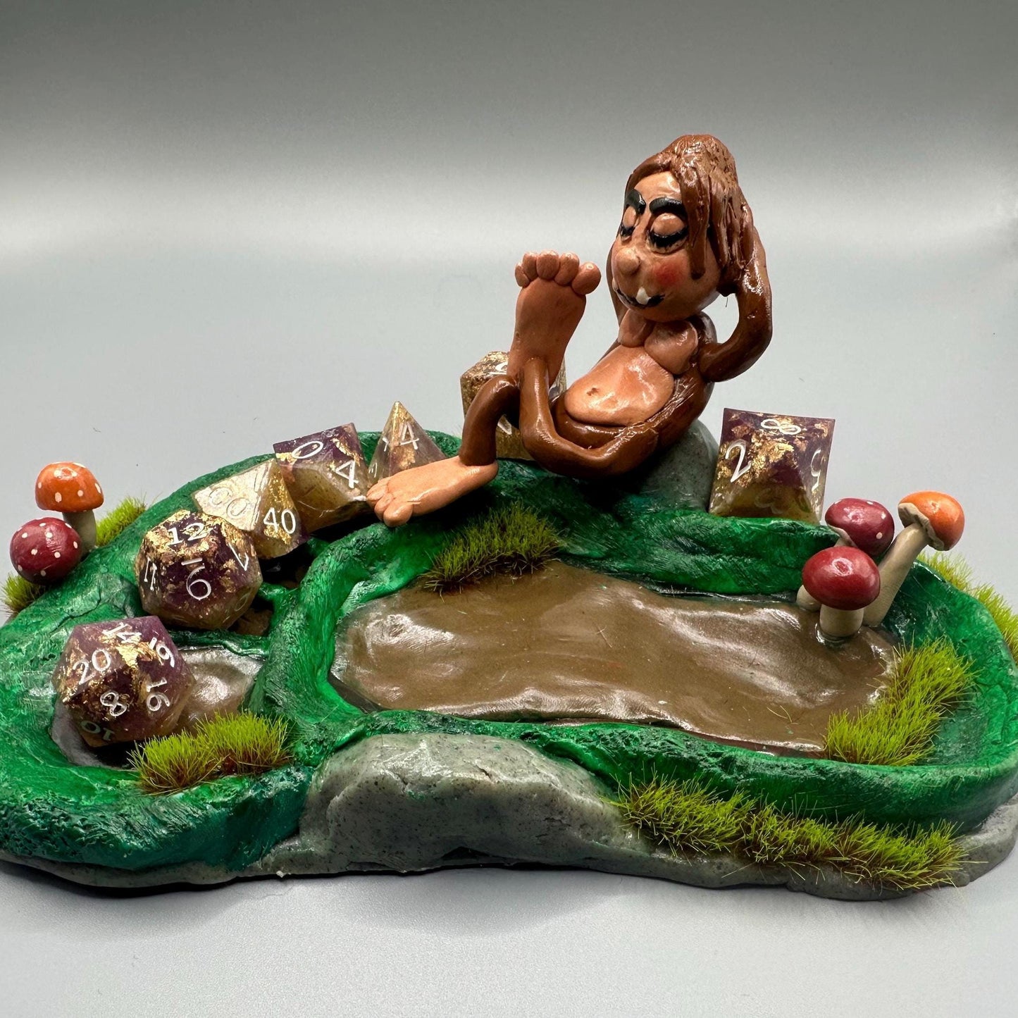Cute handmade polymer clay dice tray and holder for a cryptid lover with a baby Bigfoot theme. Perfect gift for RPG, D&D, cryptid lovers and fantasy lovers. Baby Bigfoot sitting at the edge of a full grown Bigfoot foot print in the woods surrounded by fake grass and clay mushrooms. No AI used in the making of this dice tray!