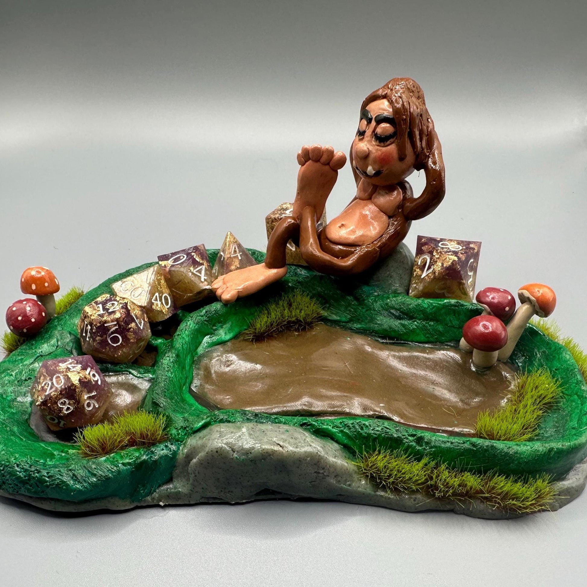 Cute handmade polymer clay dice tray and holder for a cryptid lover with a baby Bigfoot theme. Perfect gift for RPG, D&D, cryptid lovers and fantasy lovers. Baby Bigfoot sitting at the edge of a full grown Bigfoot foot print in the woods surrounded by fake grass and clay mushrooms. No AI used in the making of this dice tray!