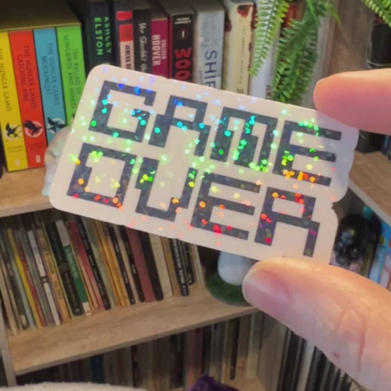 Holographic black and white gamer sticker that says GAME OVER in pixelated retro font. Water-resistant vinyl decal for laptops, bottles, consoles.
