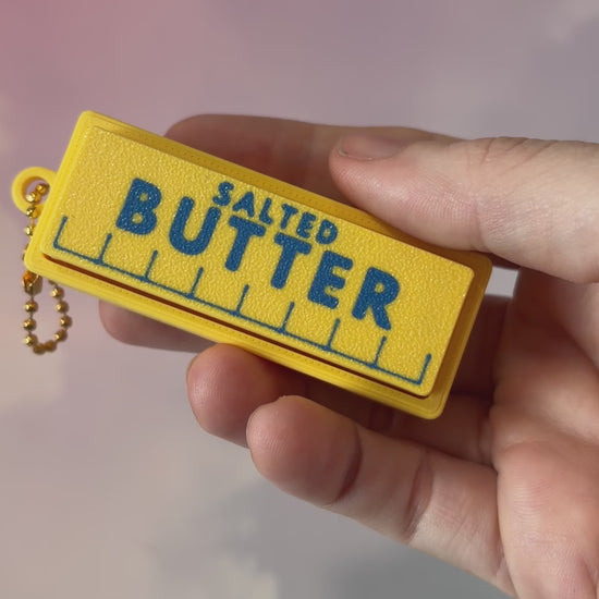 3D printed stick of butter fidget clicker keychain with classic tablespoon marking lines, gold-tone hardware, and two satisfying clicky key switches, perfect for foodies and whimsical accessory lovers.