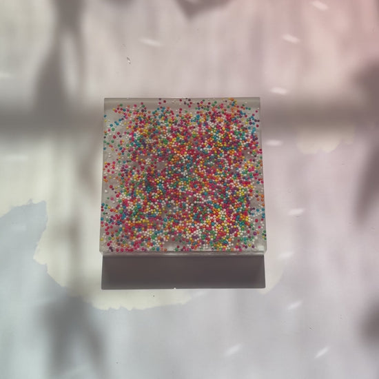 Resin coaster inlaid with rainbow sprinkles and white backing.
