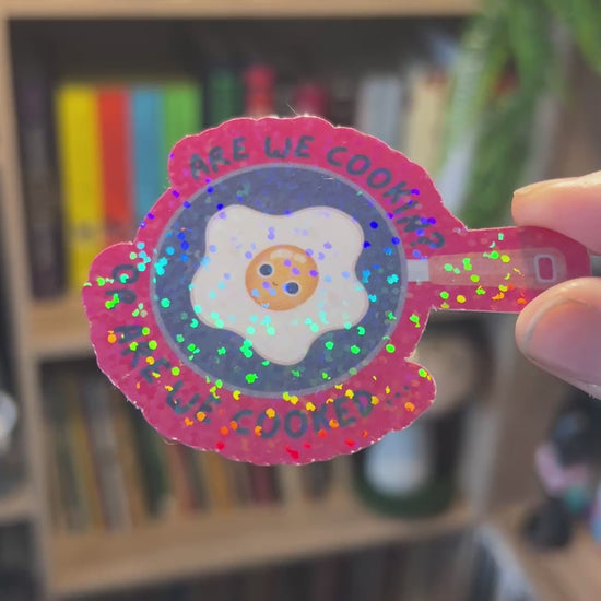Holographic vinyl sticker with a wide-eyed egg in a frying pan and the phrase “Are We Cooking or Are We Cooked?” Funny kitchen-themed design.