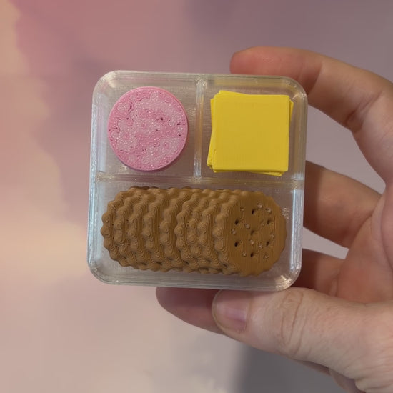 3D printed mini snack box fidget clicker with ham, cheese, and crackers under a semi-transparent lid. Ham and cheese have soft silent presses, while the crackers make satisfying audible clicks.