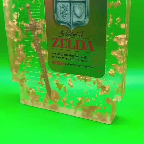 Legend of Zelda resin art featuring a retro NES cartridge with a gold sword design and gold flakes embedded. This handmade display is perfect for gamers and video game collectors, offering a unique décor piece for any gaming setup or retro collection
