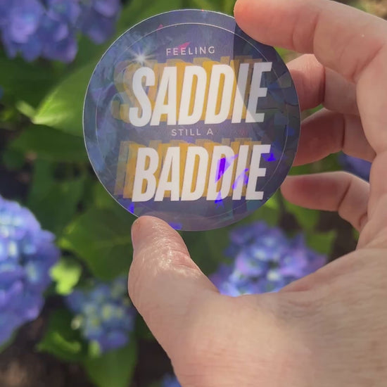Round holographic sticker with the phrase "Feeling Saddie, Still a Baddie" held in a hand with blue hydrangeas in the background.