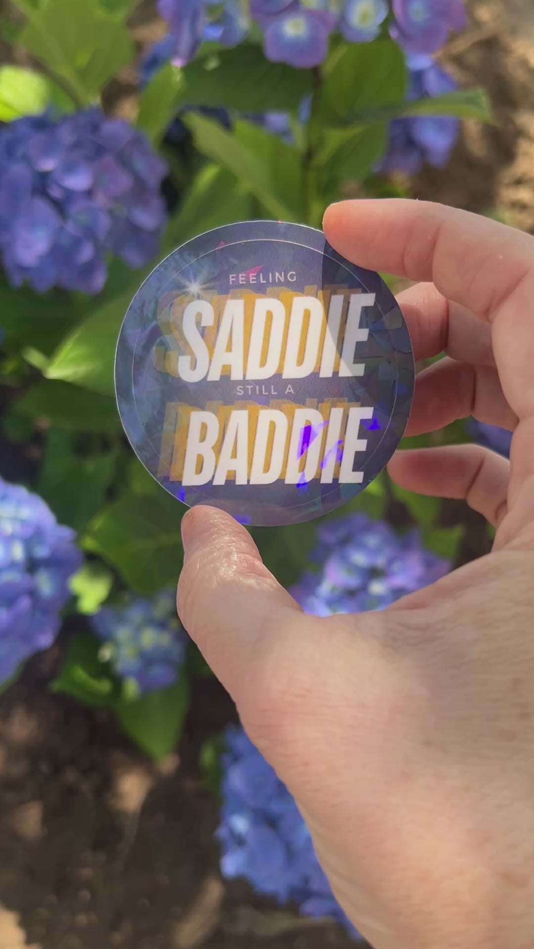 Round holographic sticker with the phrase "Feeling Saddie, Still a Baddie" held in a hand with blue hydrangeas in the background.