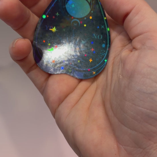 Black holographic Ouija planchette sticker with celestial sun, moon, stars, and blue-purple gradient artwork on water-resistant vinyl.