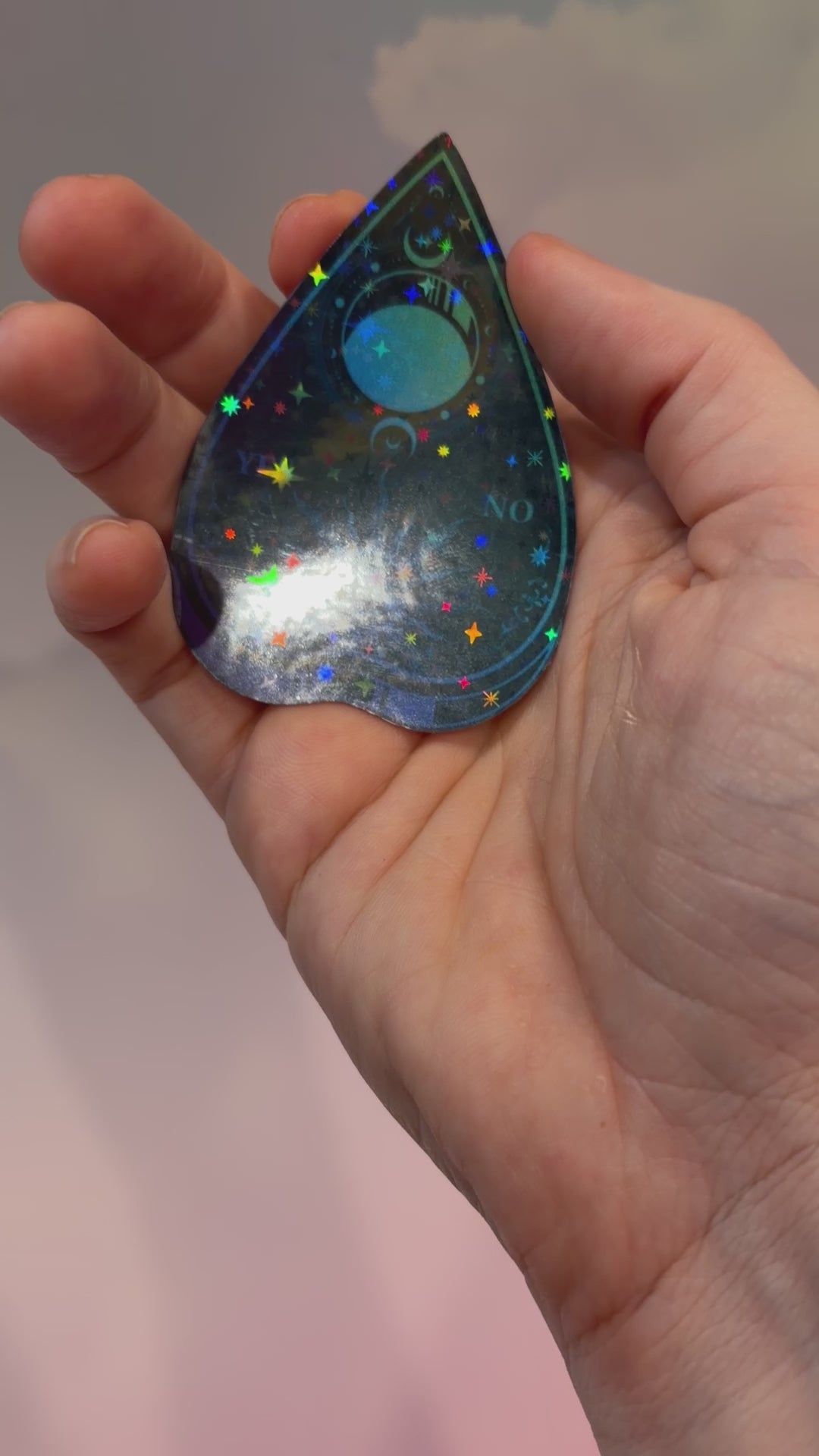 Black holographic Ouija planchette sticker with celestial sun, moon, stars, and blue-purple gradient artwork on water-resistant vinyl.