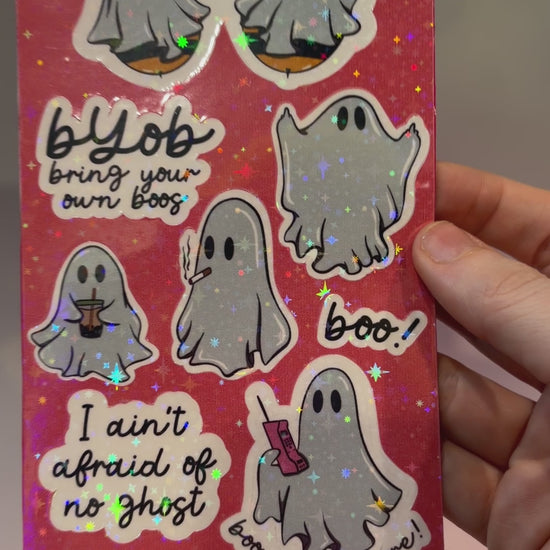 Holographic sticker sheet featuring eight cute ghost vinyl stickers with funny sayings including "Boo Bees," "BYOB Bring Your Own Boos," "I Ain't Afraid of No Ghost," and other snarky spooky designs on a pink glitter background.