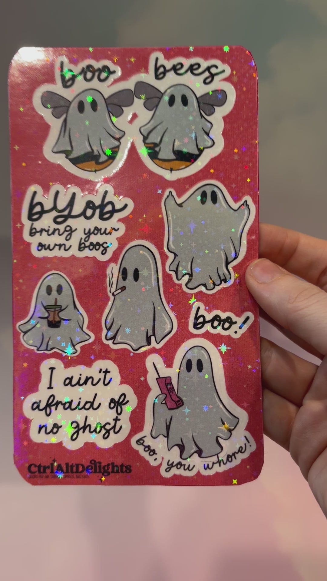 Holographic sticker sheet featuring eight cute ghost vinyl stickers with funny sayings including "Boo Bees," "BYOB Bring Your Own Boos," "I Ain't Afraid of No Ghost," and other snarky spooky designs on a pink glitter background.