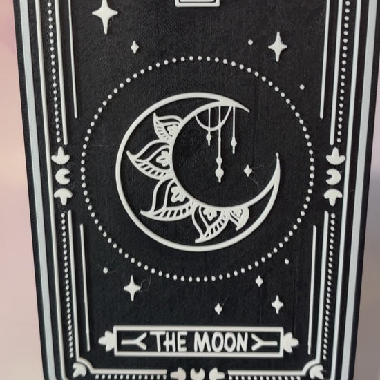 A vertical 3D printed tarot card sign featuring The Moon from the Major Arcana, displayed upright on a small black easel against a soft pink, peach, and lavender background. All 22 Major Arcana tarot card designs are available, allowing customers to choose a card that reflects their interests, spiritual practice, or personal journey.