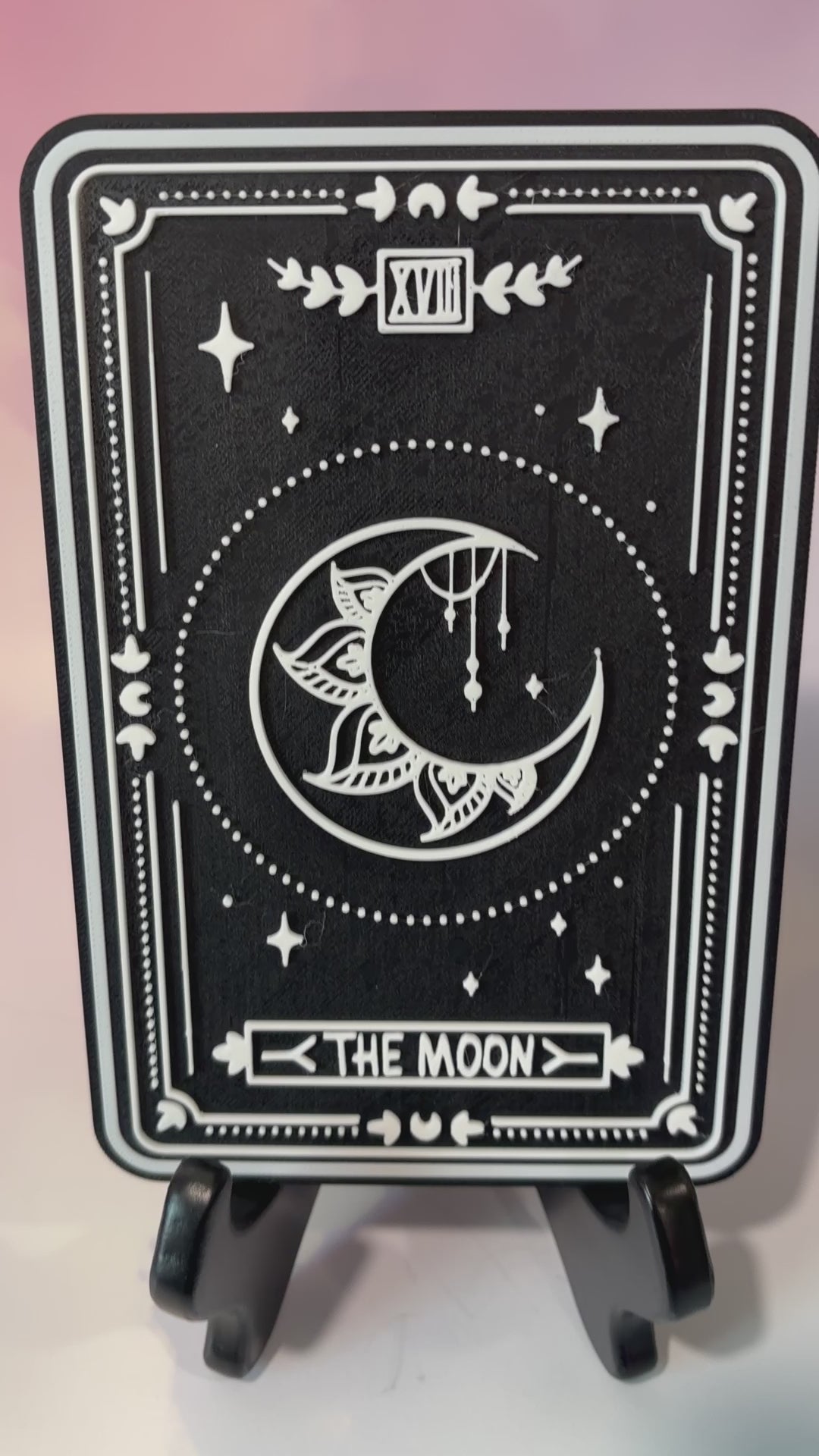 A vertical 3D printed tarot card sign featuring The Moon from the Major Arcana, displayed upright on a small black easel against a soft pink, peach, and lavender background. All 22 Major Arcana tarot card designs are available, allowing customers to choose a card that reflects their interests, spiritual practice, or personal journey.