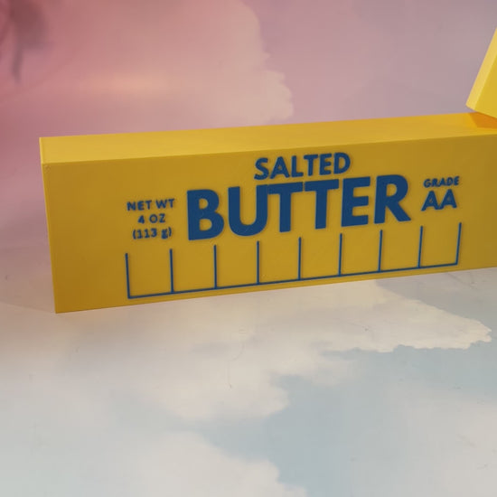 Oversized 3D printed novelty stick of butter, approximately 8.5 inches long, featuring classic tablespoon measurement lines, Grade AA labeling, and net weight details. Whimsical food-themed decor for kitchens and farmhouse-style homes.