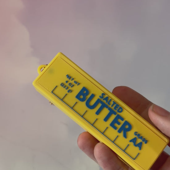 3D printed stick of butter bag charm with gold hardware, classic TBSP markings, Grade AA label, and net weight details. Sliding top hides a lipstick or chapstick in a magnetic compartment.