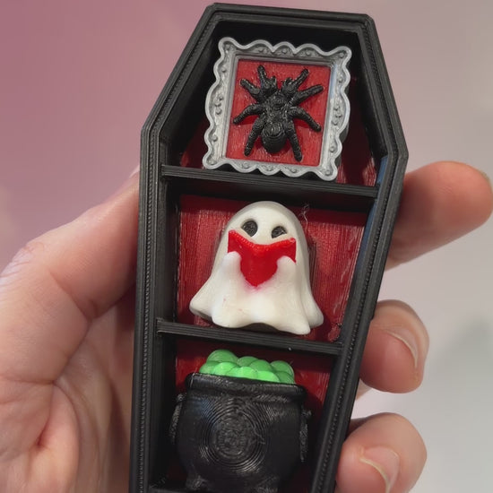 Mini 3D printed black coffin shelf fidget clicker with silver gothic trim and red interior. Features three decorative keycaps: a framed black spider, a ghost reading a book, and a bubbling green cauldron. Includes a silver keychain and is available with clicky or silent switches.