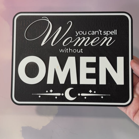 A rectangular 3D printed decorative sign featuring the phrase “You Can’t Spell Women Without Omen” in raised white lettering against a black background.