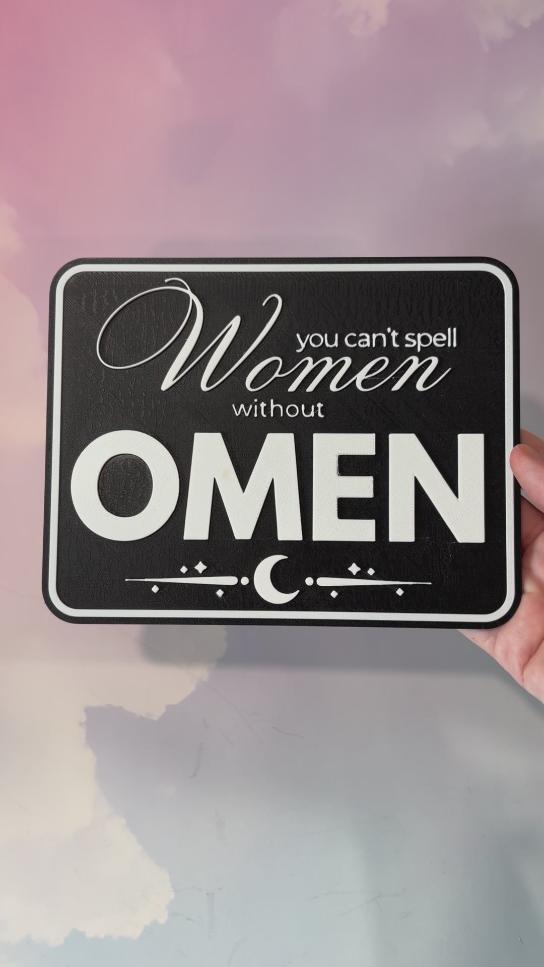 A rectangular 3D printed decorative sign featuring the phrase “You Can’t Spell Women Without Omen” in raised white lettering against a black background.