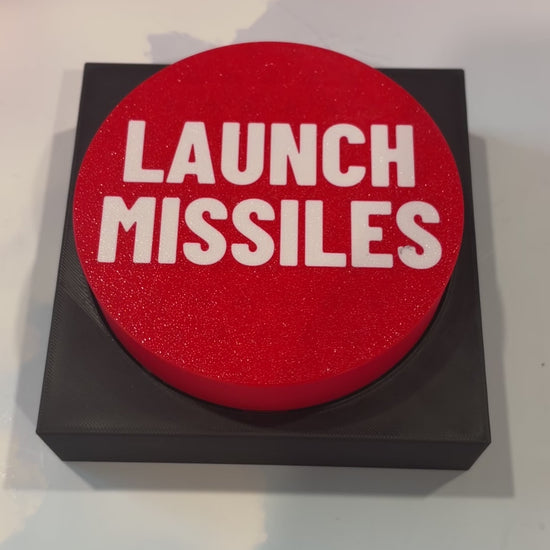Oversized red 3D printed fidget button with white “LAUNCH MISSILES” lettering on a black square base, shown on a white surface.